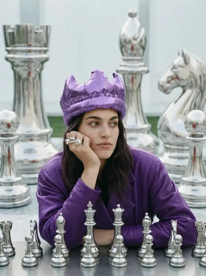 Person wearing a purple crown resting their chin beside giant silver chess pieces